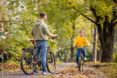 Happy father and daughter ride bicycles in autumn park on sunny day.