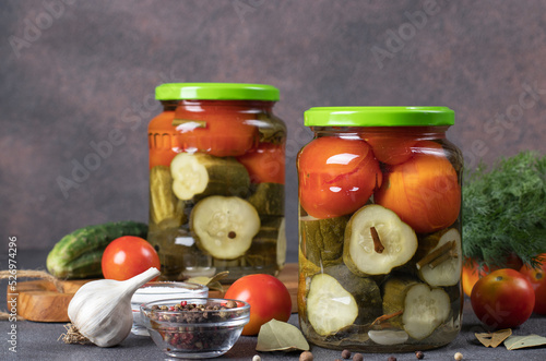 Pickled cucumbers with cherry tomatoes in two glass jars on brown background with fresh ingredients