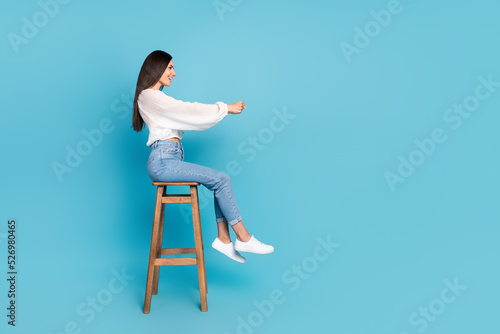 Full size profile photo of positive pretty girl sit stool arms hold imagine wheel look empty space isolated on blue color background