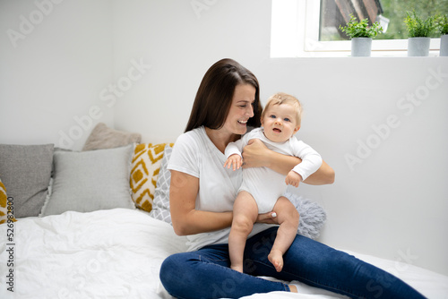 pretty young and happy mother with dark hair, white t-shirt, blue pants is cuddling in bed with her 7 months old son with blue eyes