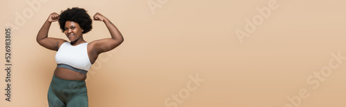 cheerful african american plus size woman in crop top showing power gesture isolated on beige, banner. © LIGHTFIELD STUDIOS