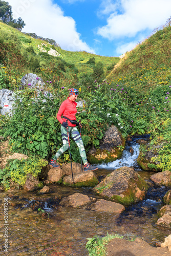 a woman in bright sports hiking clothes with trekking poles crosses a mountain stream in summer and walks along a hiking trail
