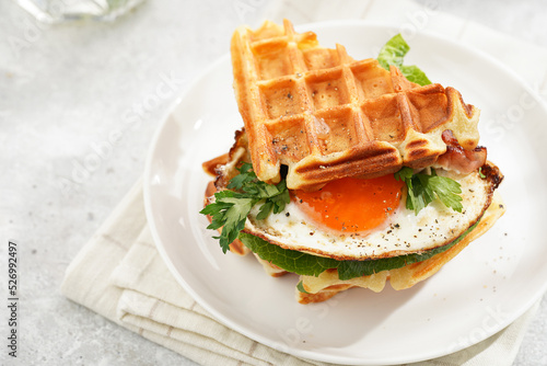 Savory breakfast - fresh homemade belgian waffle sandwich with green lettuce, crisp bacon and fried egg on white plate on beige colored table cloth, glass with lemon water, cup of coffee