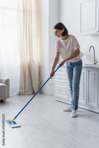 Brunette woman in casual clothes cleaning floor with mop in kitchen.