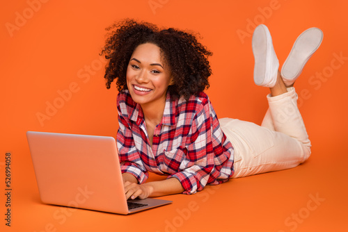 Portrait of attractive cheery girl lying using laptop ui web study education isolated on bright orange color background © deagreez