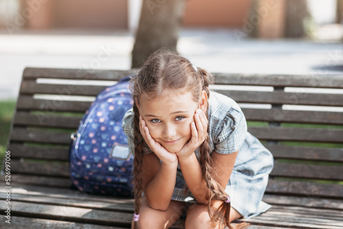 Back to school. A cute little schoolgirl in a dress with pigtails and large blue backpackis sitting on a bench in the school yard . A little girl is going to the first grade.