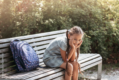Back to school. A cute little schoolgirl in a dress with pigtails and large blue backpackis sitting on a bench in the school yard . A little girl is going to the first grade. photo