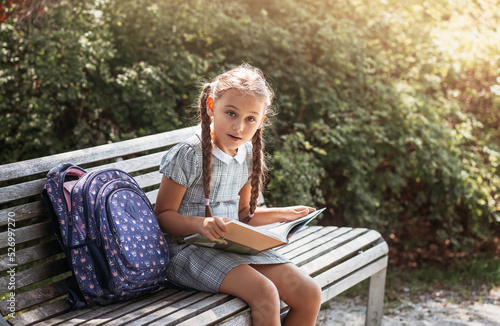 Girl with a backpack sitting on a bench and reading a book near the school. Back to school, lesson schedule, a diary with grades. Education, primary school classes, Back to school