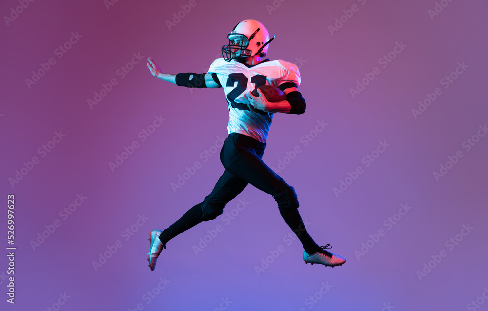 Portrait of sportive man, american football player in uniform playing, training isolated over purple background in neon light.