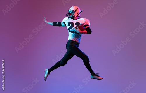 Portrait of sportive man, american football player in uniform playing, training isolated over purple background in neon light.