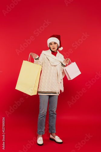 full length of surprised young woman in santa hat and winter outfit holding shopping bags on red.