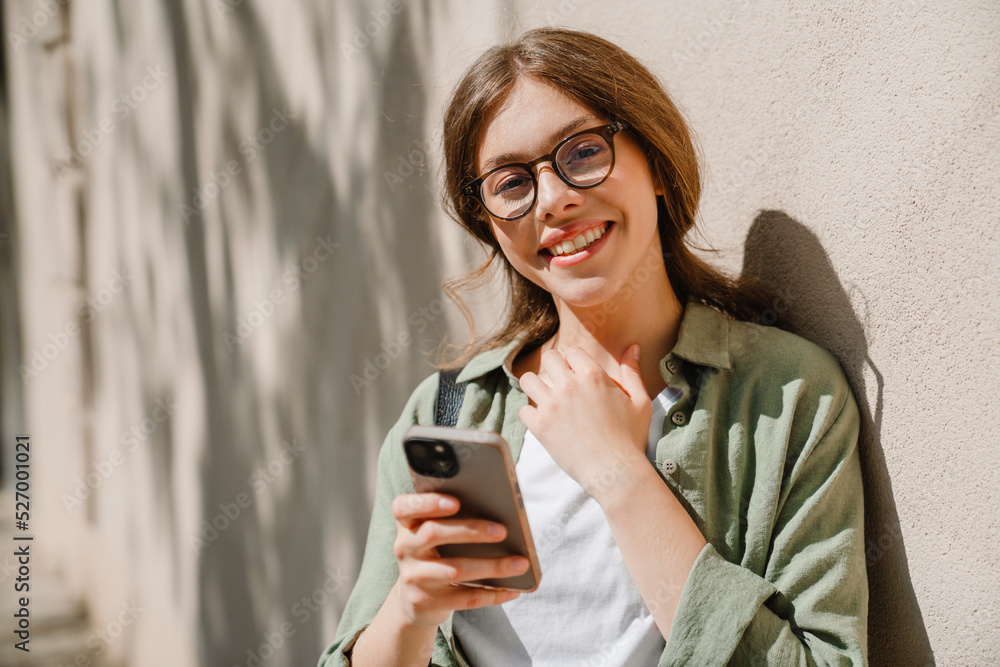 Fototapeta premium Young beautiful smiling woman in glasses holding phone touching neck