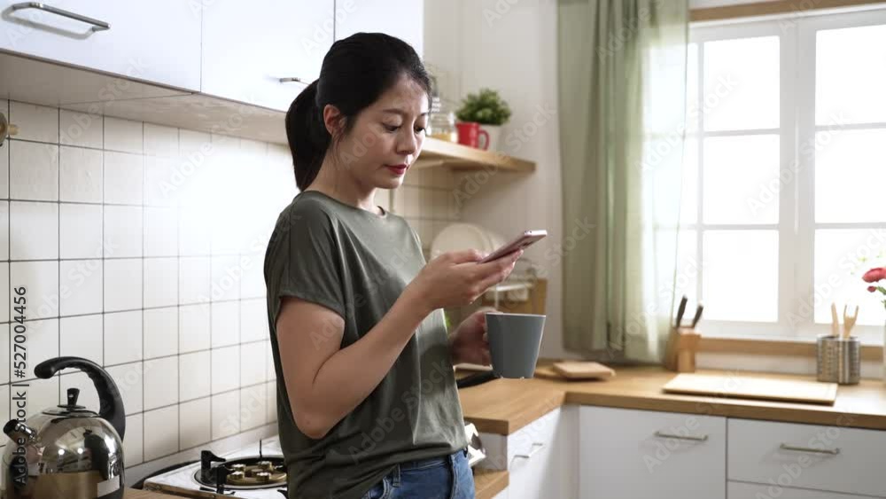 portrait of an asian female holding tea cup and checking messages on the mobile phone in a modern kitchen with window and daylight at home. portrait of an asian female holding tea cup and checking messages on the mobile phone in a modern kitchen with window and daylight at home.