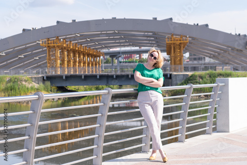 A portrait of a girl in a green blouse, trousers and sunglasses against background of unfinished urban architecture. A girl walks around a new area of the city.