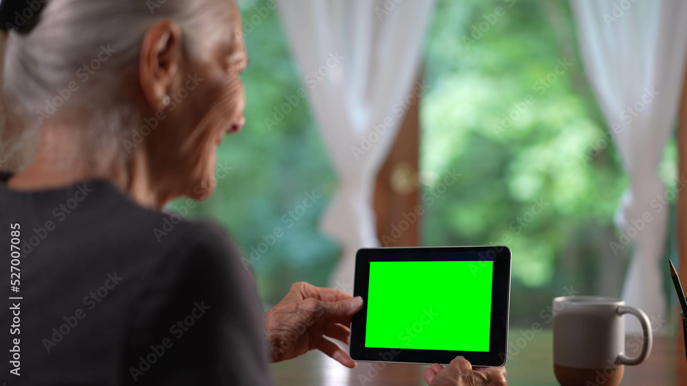 Over shoulder view of smiling senior elderly woman making video call on tablet computer with green screen talking and listening during video chat with friends or colleagues.
