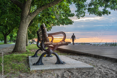 The Boardwalk on Balmy Beach in Toronto at daybreak with a park bench in the foreground and a person walking in the background.