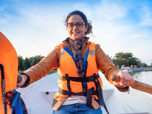 Beautiful brunette woman teen with glasses and in an orange life jacket rowing oars while sitting in a boat. Family walks at the boat station. Illuminated by sun glare