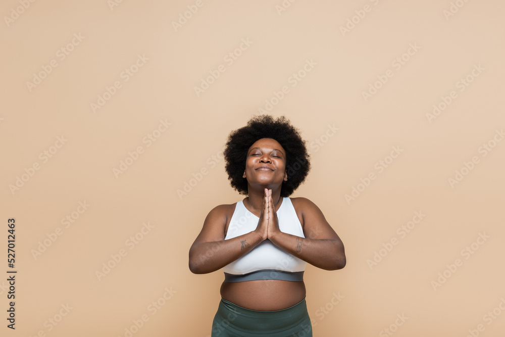 pleased african american plus size woman in crop top praying isolated on beige. pleased african american plus size woman in crop top praying isolated on beige.