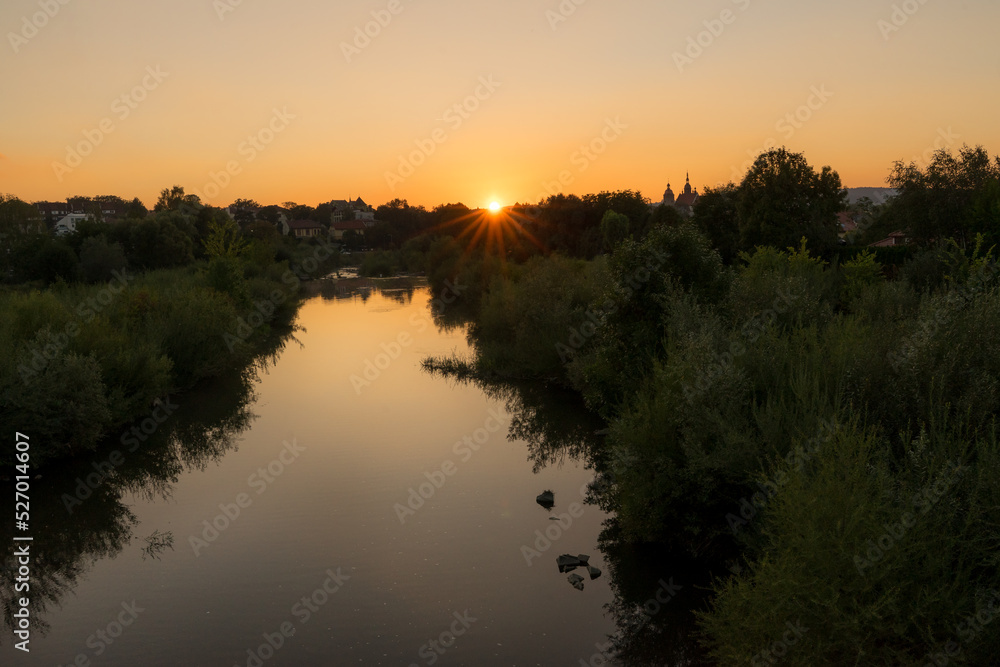 Sunset on the river surrounded by willows. Trees are reflected in still water. Building of town in the background.