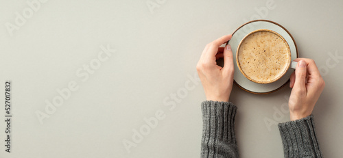 Autumn mood concept. First person top view photo of female hands in sweater holding cup of frothy coffee and saucer on isolated grey background with copyspace photo
