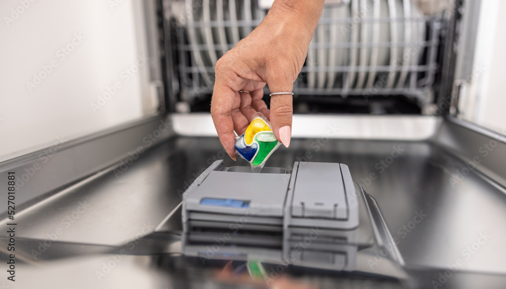 Blue, yellow and green dishwashing tablet put into the appliance by a female hand Blue, yellow and green dishwashing tablet put into the appliance by a female hand