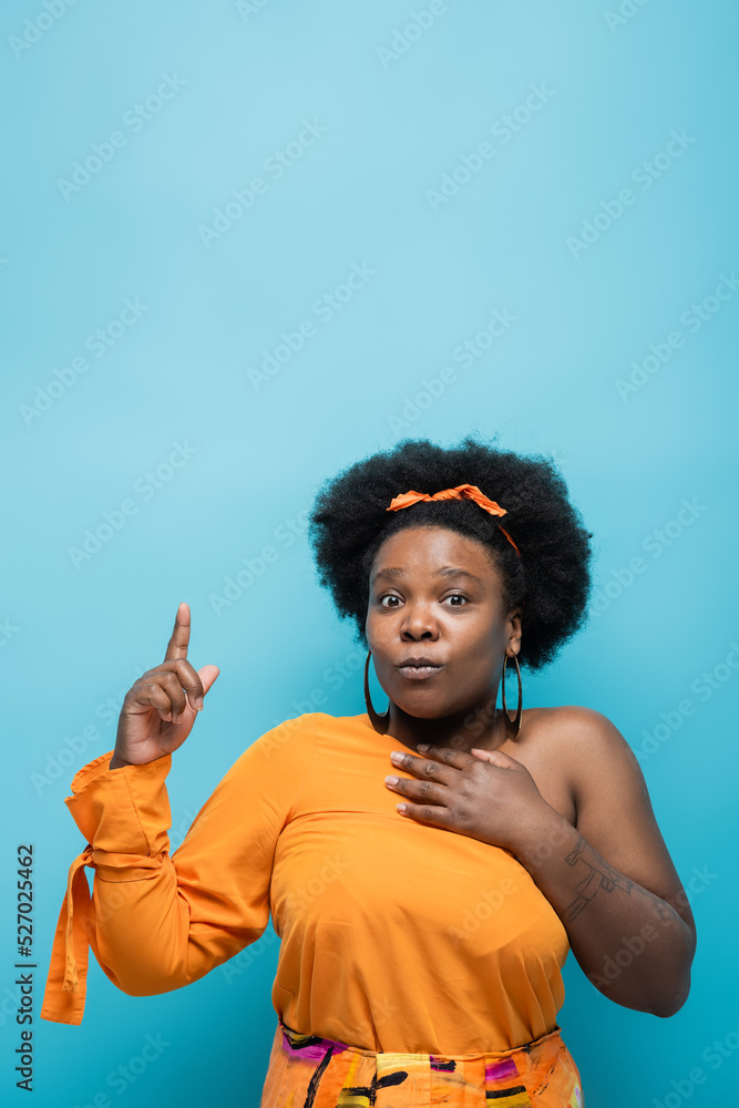 tattooed african american body positive woman in orange dress and hoop earrings pointing with finger isolated on blue. tattooed african american body positive woman in orange dress and hoop earrings pointing with finger isolated on blue.