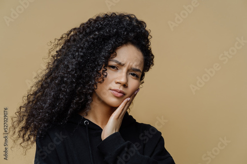 Headshot of displeased curly haired woman suffers from toothache, touches cheek, needs to see dentist, wears black sweatshirt, isolated on beige background. Tooth pain and dentristy concept. photo