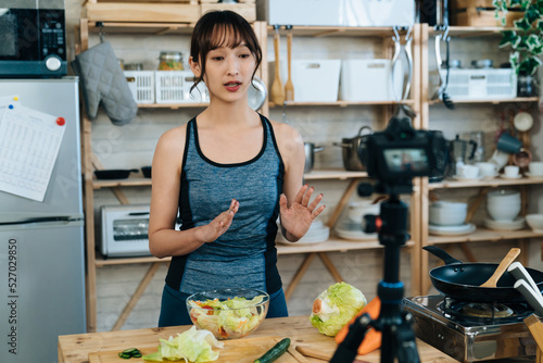 Asian fitness influencer is explaining with hand gestures while recording a video about salad making with digital camera on tripod in the kitchen at home. photo