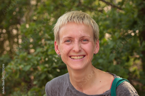 Young woman with a short hairstyle with a nose piercing laughs joyfully in the forest  on green trees background 
