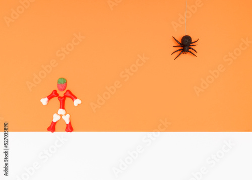 Representation of a man made of jelly beans and a black toy spider for Halloween. White and orange background.