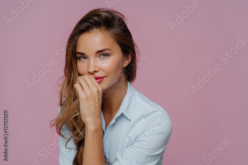 Photo of self assured attractive woman with long hair, keeps hand on lips, dressed in blue shirt, looks at camera with blue eyes, wears makeup, poses against purple background. Human face expressions
