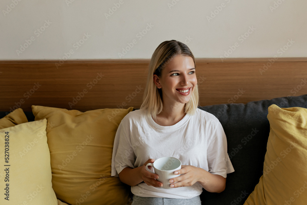 Smiling adorable lady with blond hair sitting on the coach and smiling at window in. sunny morning . Happy stylish girl start new day at home Smiling adorable lady with blond hair sitting on the coach and smiling at window in. sunny morning . Happy stylish girl start new day at home