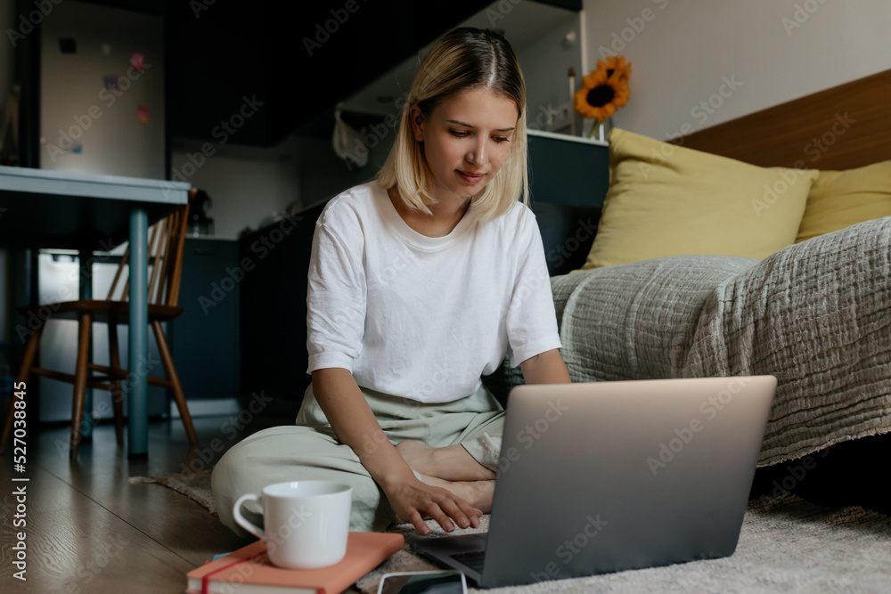 Fototapeta premium Blond woman wearing white t-shirt and light pants sitting on the floor at home and working on laptop with coffee. Adorable girl working remote at home