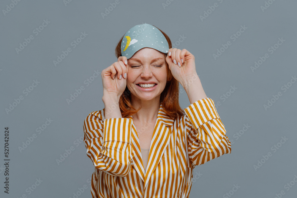 Good morning, lifestyle. Smiling freckled woman takes off blindfold, wears yellow striped pajama, awakes with smile after seeing pleasant dreams, feels relaxed and refreshed, poses over grey wall