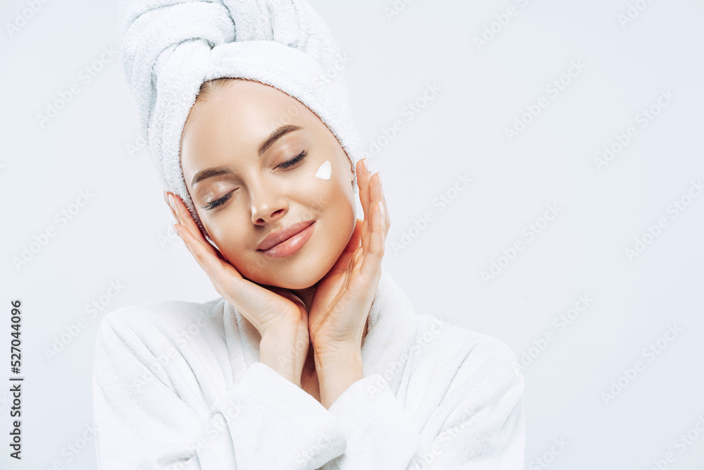Studio shot of relaxed beautiful woman with closed eyes tilts head, touches skin gently, applies face cream, wears bath towel on head after taking spa procedures, enjoys facial treatments, body care Studio shot of relaxed beautiful woman with closed eyes tilts head, touches skin gently, applies face cream, wears bath towel on head after taking spa procedures, enjoys facial treatments, body care