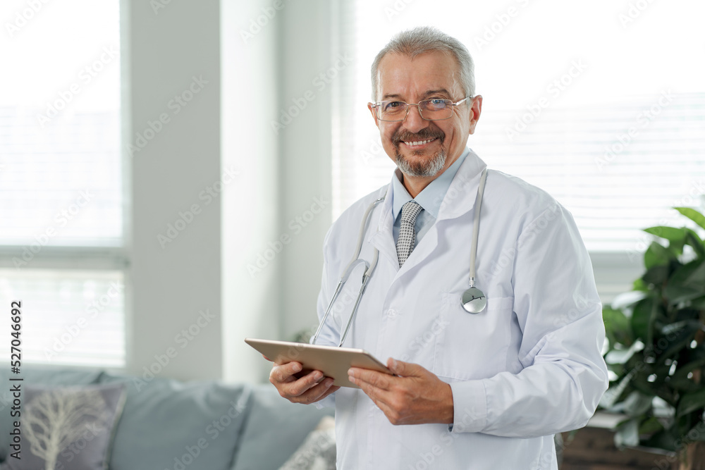 Portrait of an elderly doctor, he looks into the camera. The doctor stands against the background of a medical clinic and holds a tablet in his hands. Portrait of an elderly doctor, he looks into the camera. The doctor stands against the background of a medical clinic and holds a tablet in his hands.
