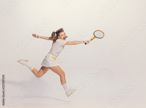Portrait of young emotive girl in white uniform playing badminton isolated over grey studio background