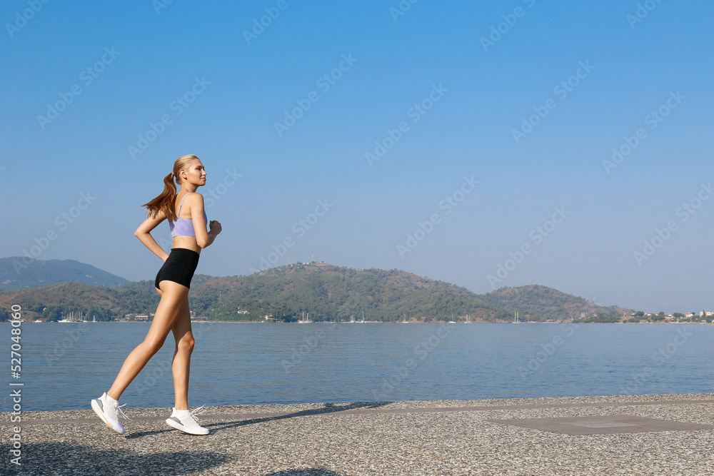 Fit blonde woman wearing black shorts and lilac tank top exercising in the park. Female model in a sportswear running along the coast on a beautiful summer day. Close up, copy space, background.