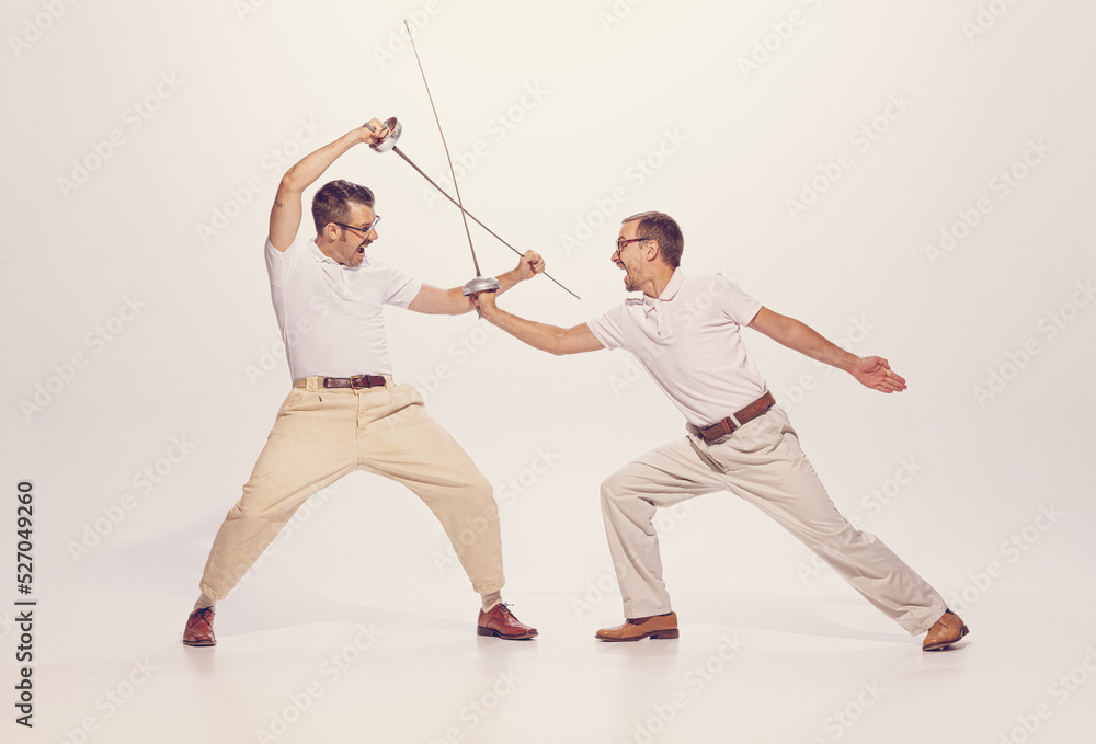 Portrait of two men in a suit fighting with swords isolated over grey studio background. Expressing emotions