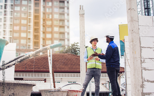Two diversity male engineers team working, inspecting outdoor at construction site, wearing hard hats for safety, talking, discussing, using tablet to plan for building. Career, Industry Concept.