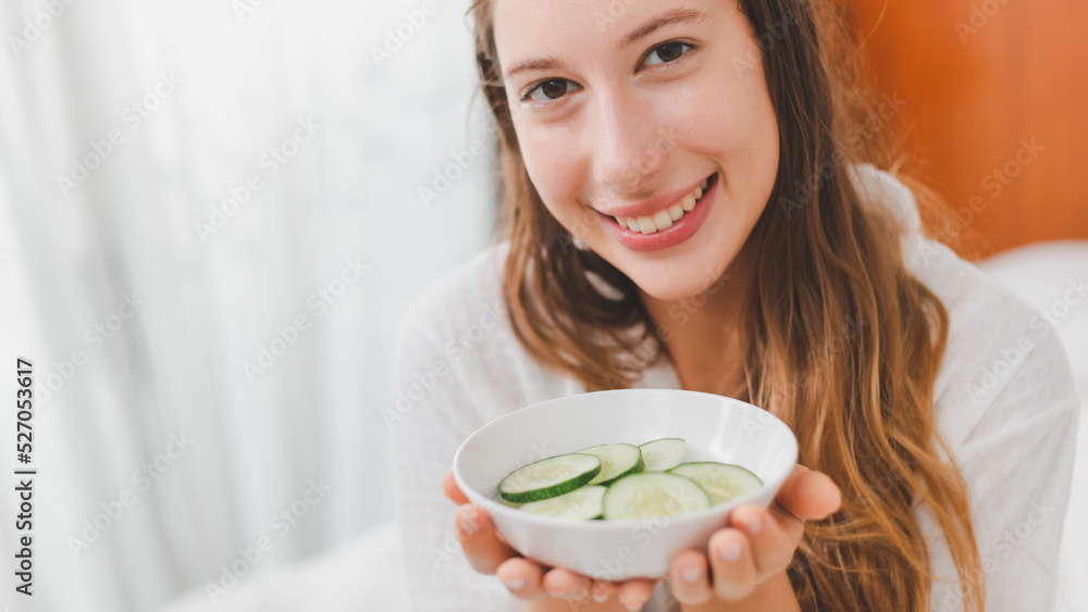 Young Makeup free face Caucasian woman holding bowl of cucumber slices preparing for beauty skin care routine at home. Attractive happy lady with bare skin face in bathrobe doing skin treatment. Young Makeup free face Caucasian woman holding bowl of cucumber slices preparing for beauty skin care routine at home. Attractive happy lady with bare skin face in bathrobe doing skin treatment.