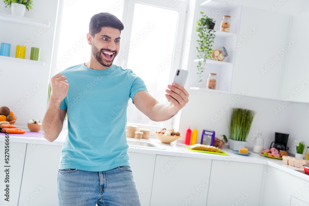 Photo of delighted overjoyed man hold telephone raise fist success accomplishment luck kitchen house indoors Photo of delighted overjoyed man hold telephone raise fist success accomplishment luck kitchen house indoors