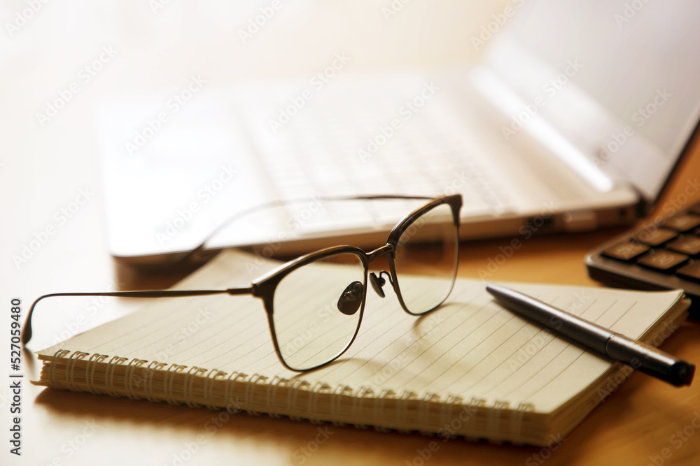 Glasses on notebook with pen and laptop on the office table; office or home working space concept Glasses on notebook with pen and laptop on the office table; office or home working space concept