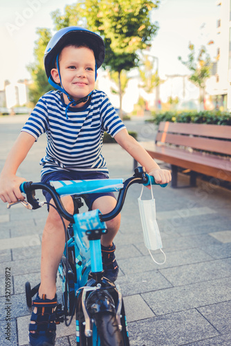 A little boy rides a bicycle on the city streets with a protective mask on the handlebars. A beautiful boy enjoying riding a bicycle.