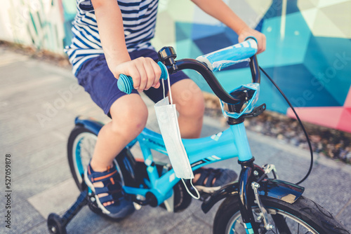 Close-up of an unrecognized boy riding a bicycle on a sunny day, his face mask hanging on the handlebars for protection.