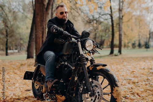 Male motorcyclist drives in nature on fast bike  wears shades  black jacket  gloves  jeans and boots  enjoys autumn season  spends free time actively  ready for long trip. People  transport  driving