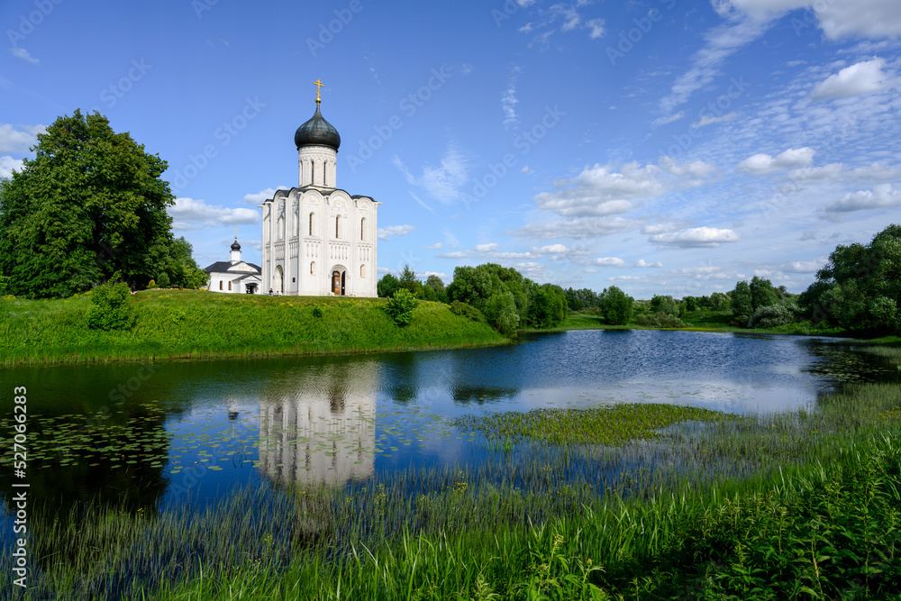 View of the church of Pokrova-na-Nerli and its reflection in the river among trees and meadows in Bogolyubovo, Russia