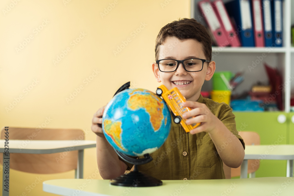 Cute caucasian schoolboy wearing glasses and sits at a desk in the classroom at elementary school. Little boy next the Globe playing with yellow school bus toy. Back to school concept Cute caucasian schoolboy wearing glasses and sits at a desk in the classroom at elementary school. Little boy next the Globe playing with yellow school bus toy. Back to school concept