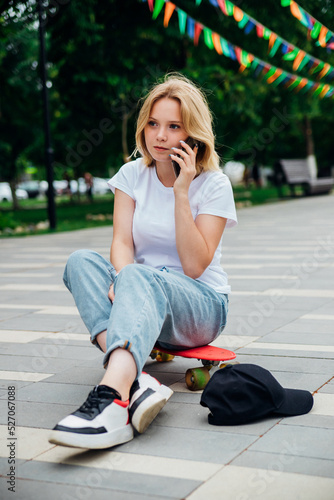 A young beautiful teen girl is sitting on a skateboard on a summer day in the park. A girl is talking on a mobile phone in the open air. Chat with friends.