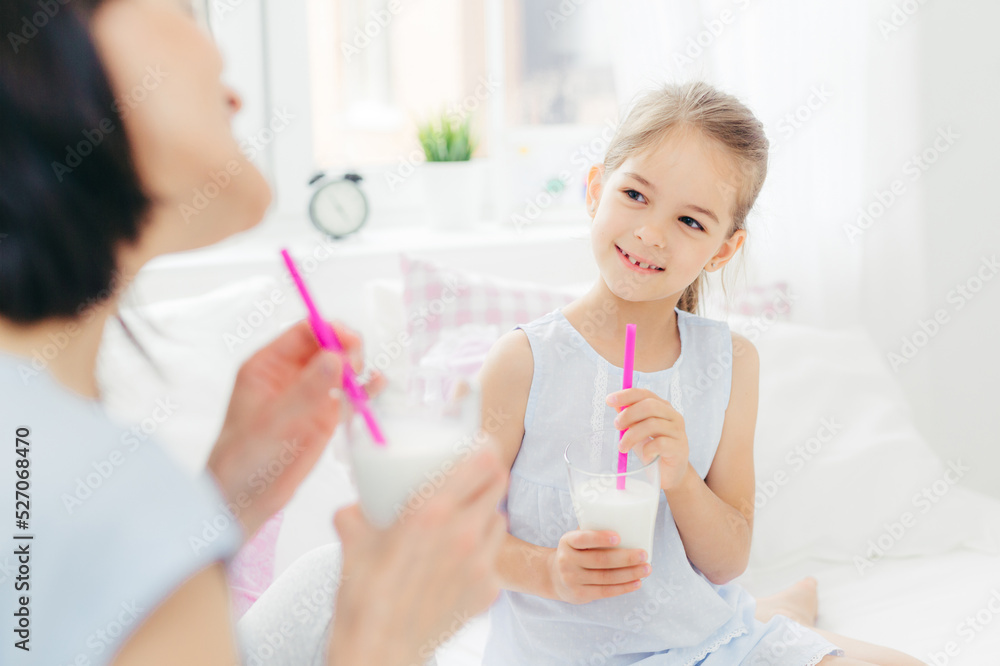Cropped shot of beautiful small female child drinks tasty milk shake together with her mother, enjoys good morning and calm domestic atmosphere, has happy look. People, breakfast and bedding Cropped shot of beautiful small female child drinks tasty milk shake together with her mother, enjoys good morning and calm domestic atmosphere, has happy look. People, breakfast and bedding